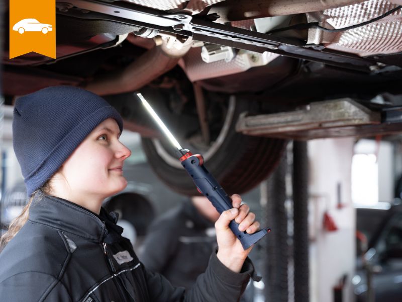 Zu sehen ist eine Frau die mit einer Leuchte unter einem Auto steht 