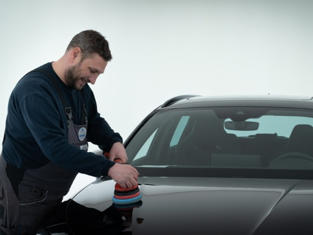Zu sehen ist ein männlicher Mitarbeiter, der gerade von einem schwarzen Fahrzeug die Motorhaube poliert
