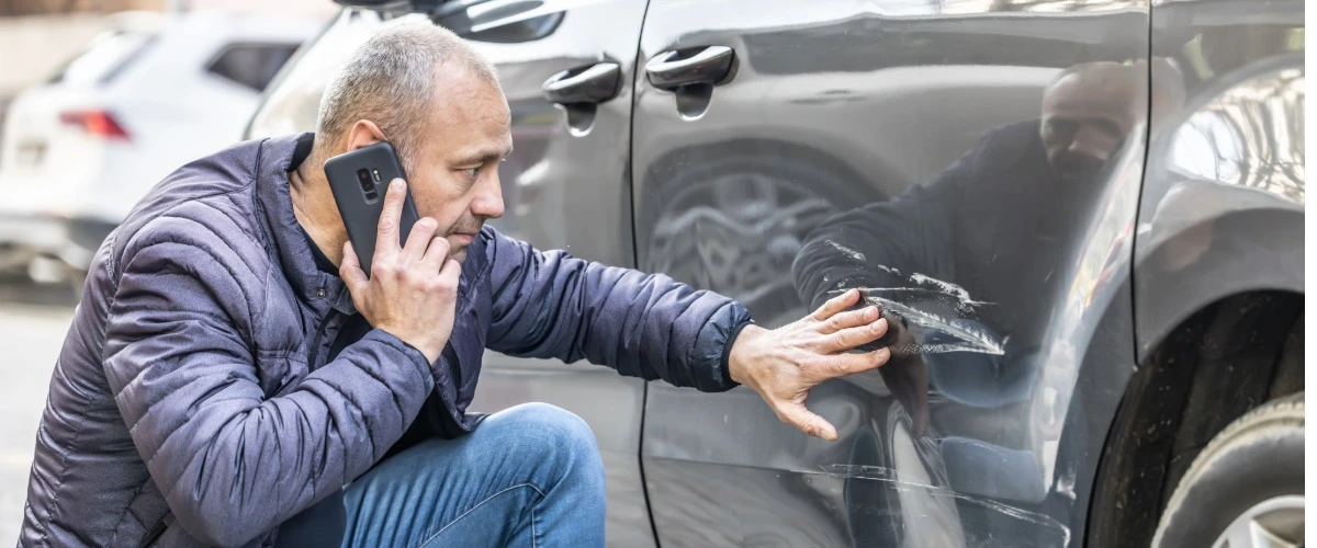Ein Mann telefoniert und kniet vor einer Autotür, die verkratzt ist.