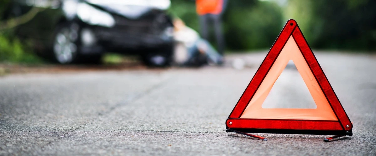 Im Vordergrund ist ein aufgestelltes Warndreieck zu sehen, im Hintergrund ein kaputtes Auto